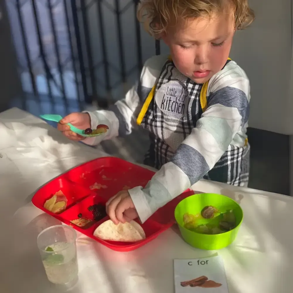 Child reaches into a red tray with food at a table, holding a spoon beside a green bowl.