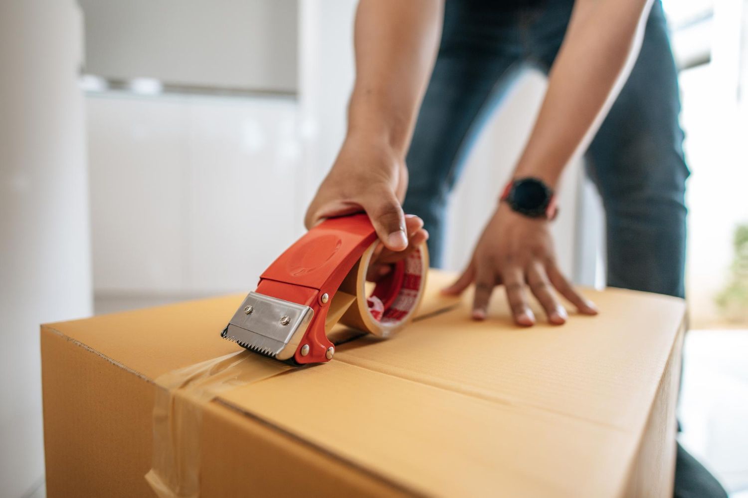 Hands sealing a cardboard box with red packing tape during moving or shipping.