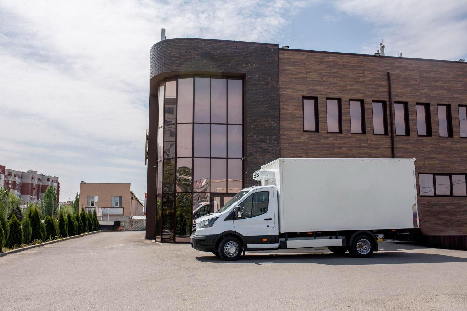 White box truck parked beside a brick building with large windows on a quiet street.