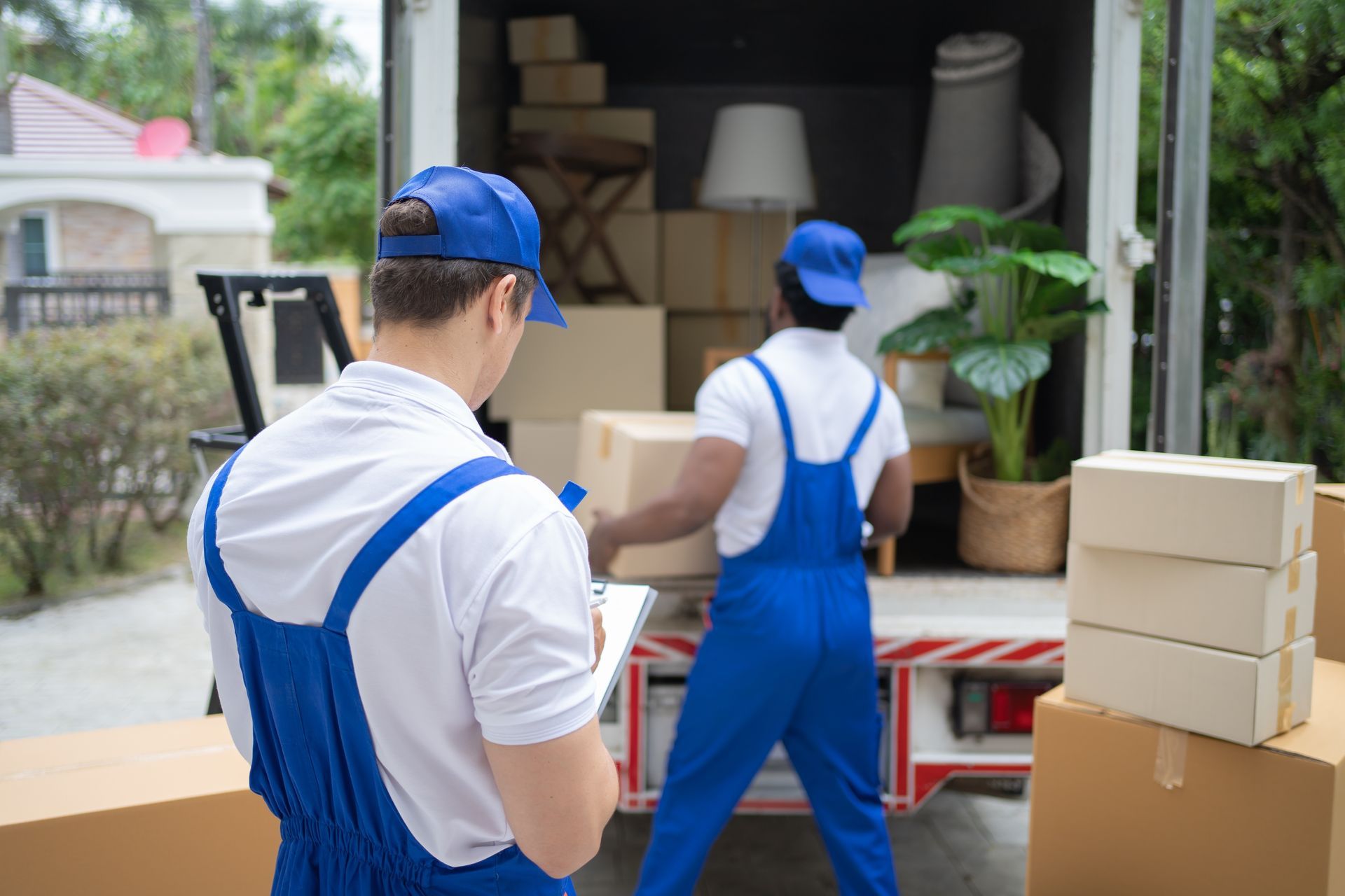 Two movers in blue overalls load boxes into a truck outside a building