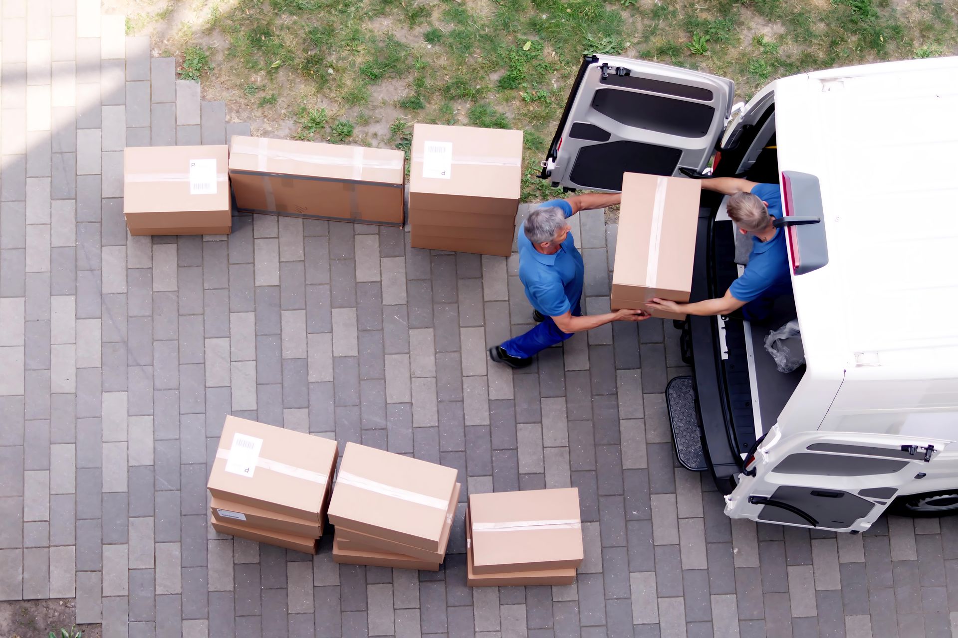 Two delivery workers unload cardboard boxes from a white van onto a paved driveway.