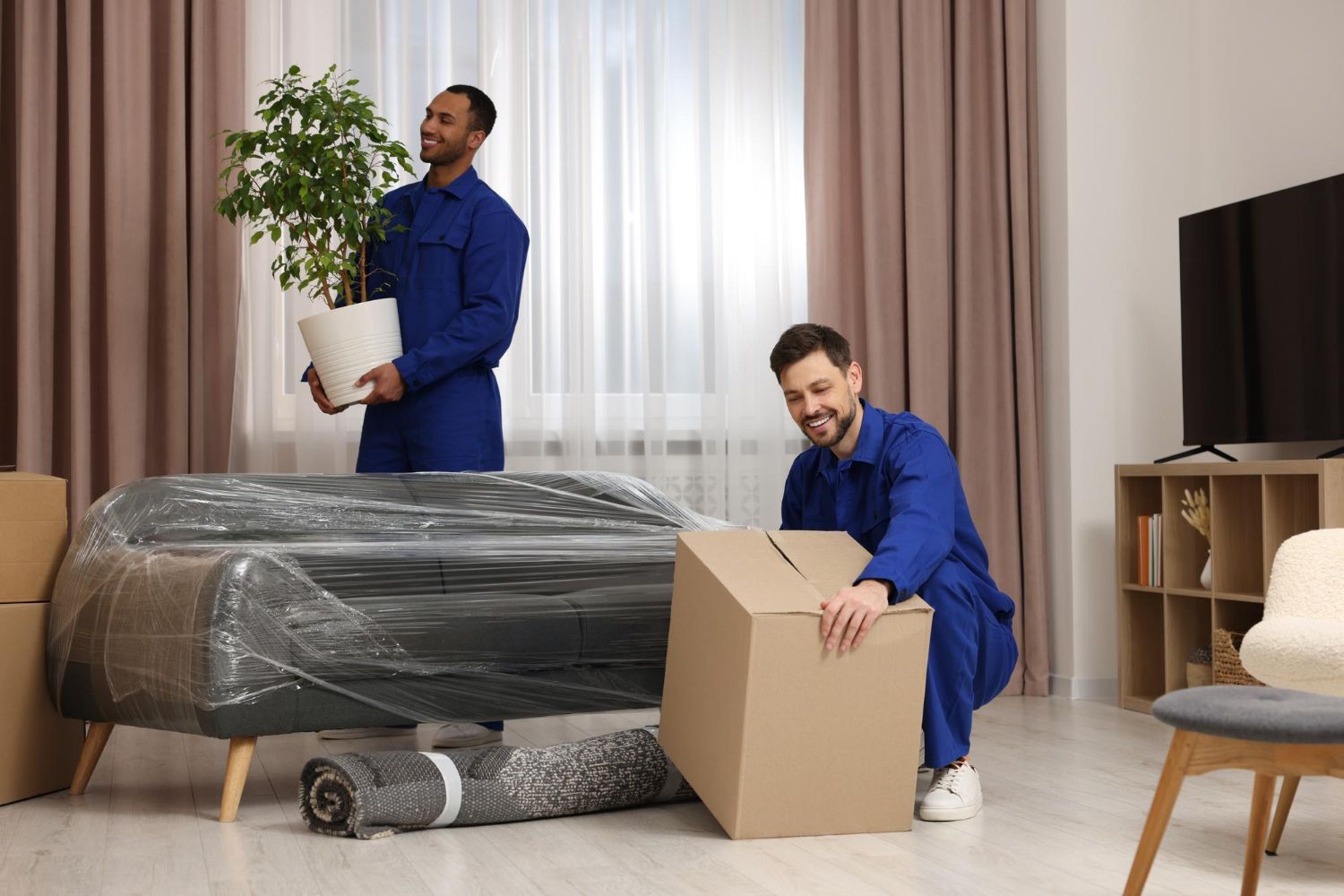 Two movers in blue uniforms unpack furniture in a living room with a sofa wrapped in plastic.