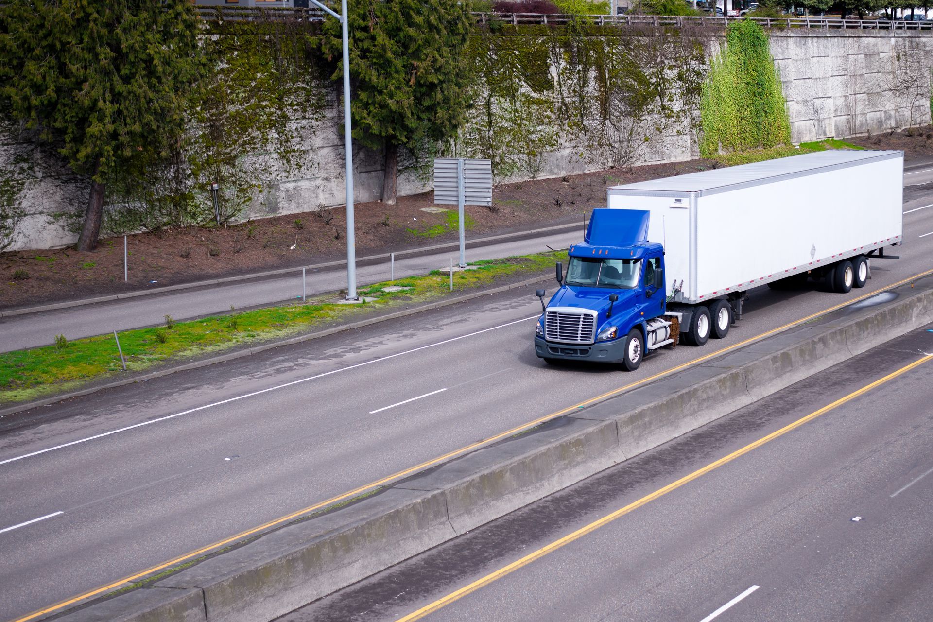 Blue semi-truck hauling a white trailer on a multi-lane road beside a stone wall and greenery