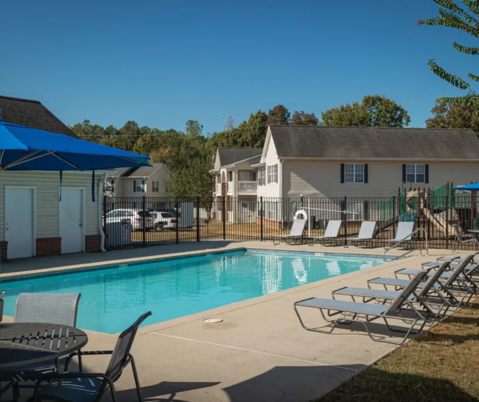 A sunny outdoor swimming pool at an apartment complex, surrounded by lounge chairs and a blue shade umbrella.