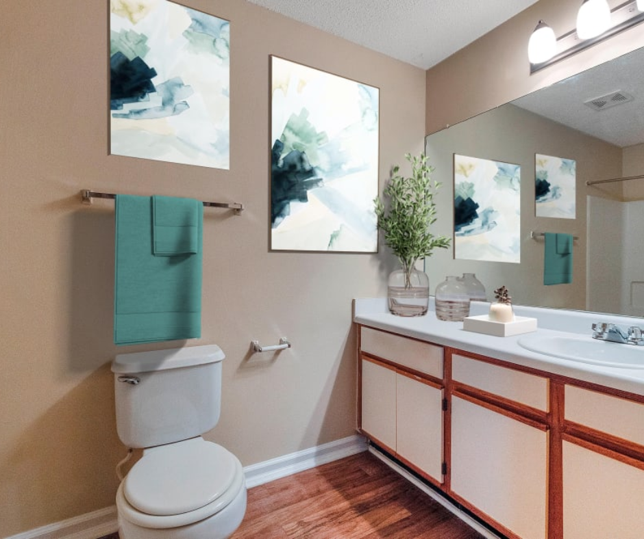 A bathroom with beige walls, teal towels, white vanity, and abstract art prints above a toilet and vanity mirror.