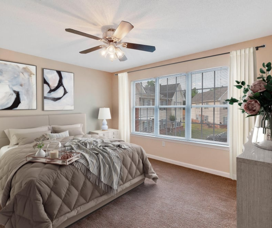 A modern bedroom features a beige bed, two abstract artworks on the wall, a ceiling fan, and large windows with curtains.