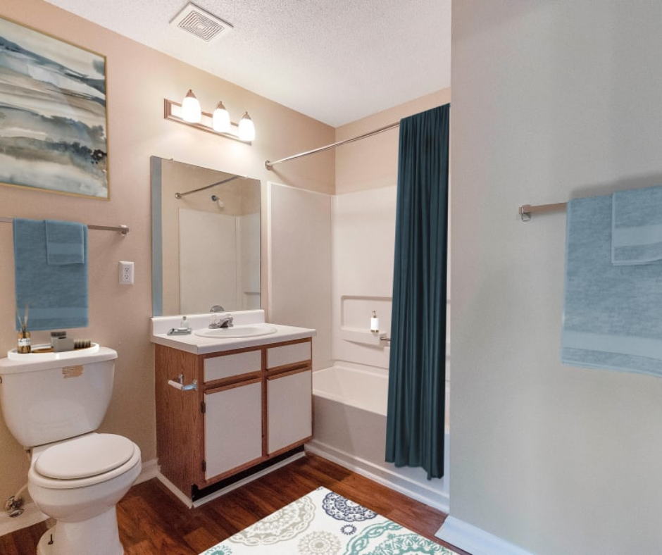 A bright bathroom with a wood vanity, white toilet, patterned rug, and a shower with a dark blue curtain.
