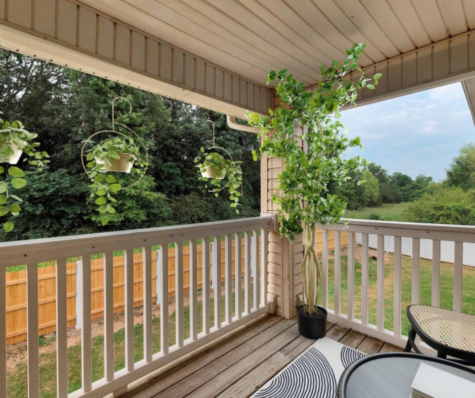 A wooden porch with a potted tree, hanging plants, a patterned rug, and a small table overlooking a grassy yard.