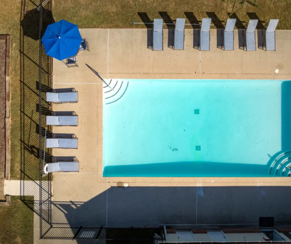 An overhead view of a rectangular swimming pool with lounge chairs, a blue umbrella, and a fence on a patio.