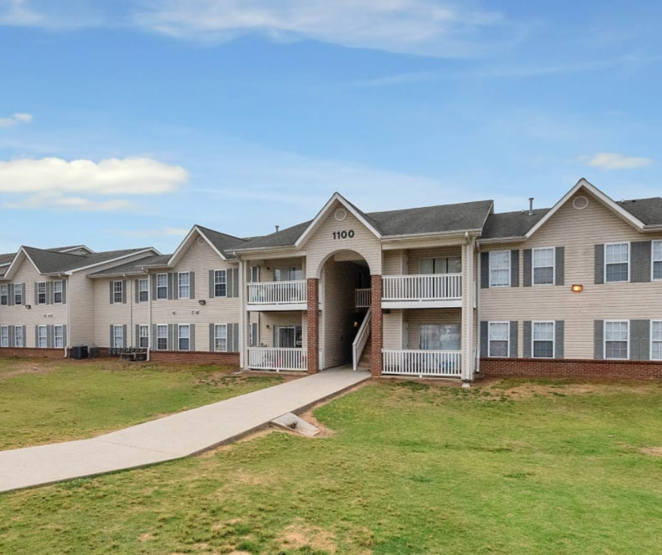 A tan, two-story apartment complex with dark shutters and a central arched entrance under a blue sky with clouds.