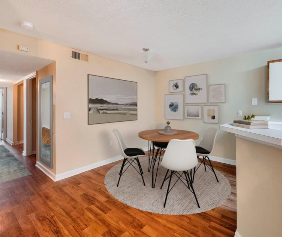 A small dining area with a round wooden table, four white modern chairs, a circular rug, and art on the walls.