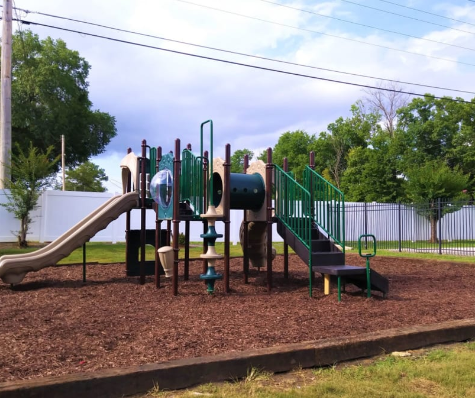 A tan and green modular playground set with a slide, climbing tube, and stairs, set in a yard covered with wood mulch.