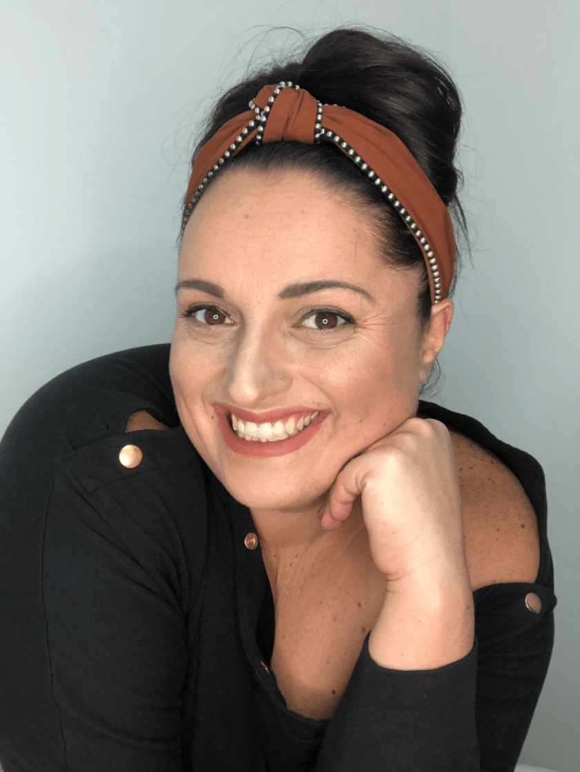 Smiling woman in a black top with a brown headband, resting her chin on her hand against a gray background