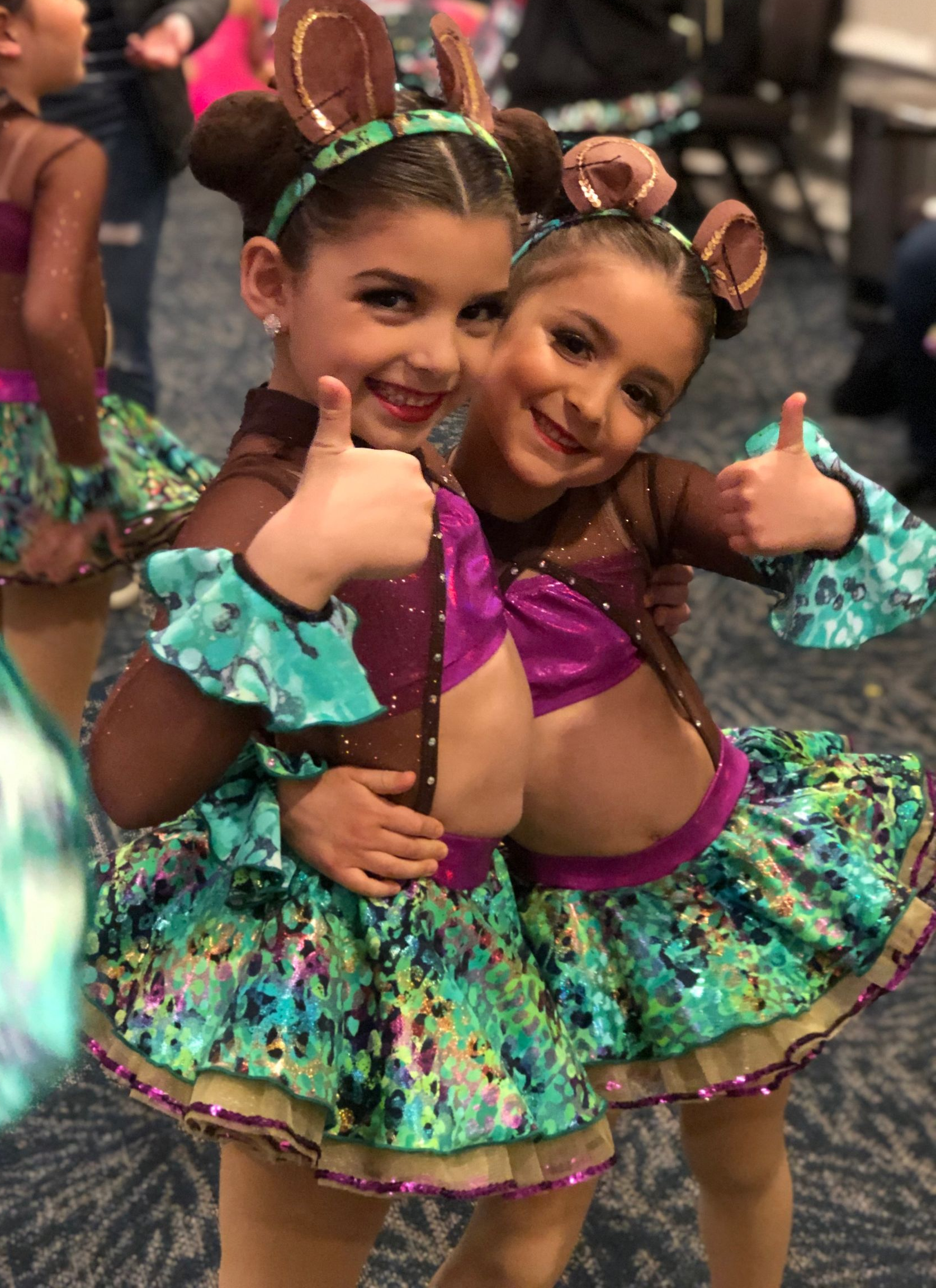 Two young dancers in colorful costumes smiling and giving thumbs up backstage