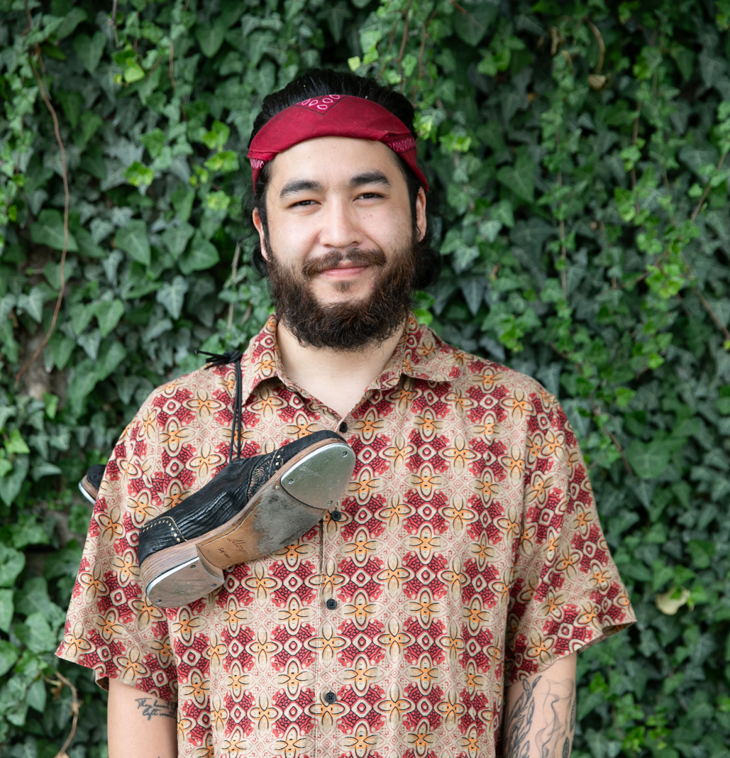 Man with beard and red bandana, shirt and light pants, tap shoe around neck, hands in pockets.