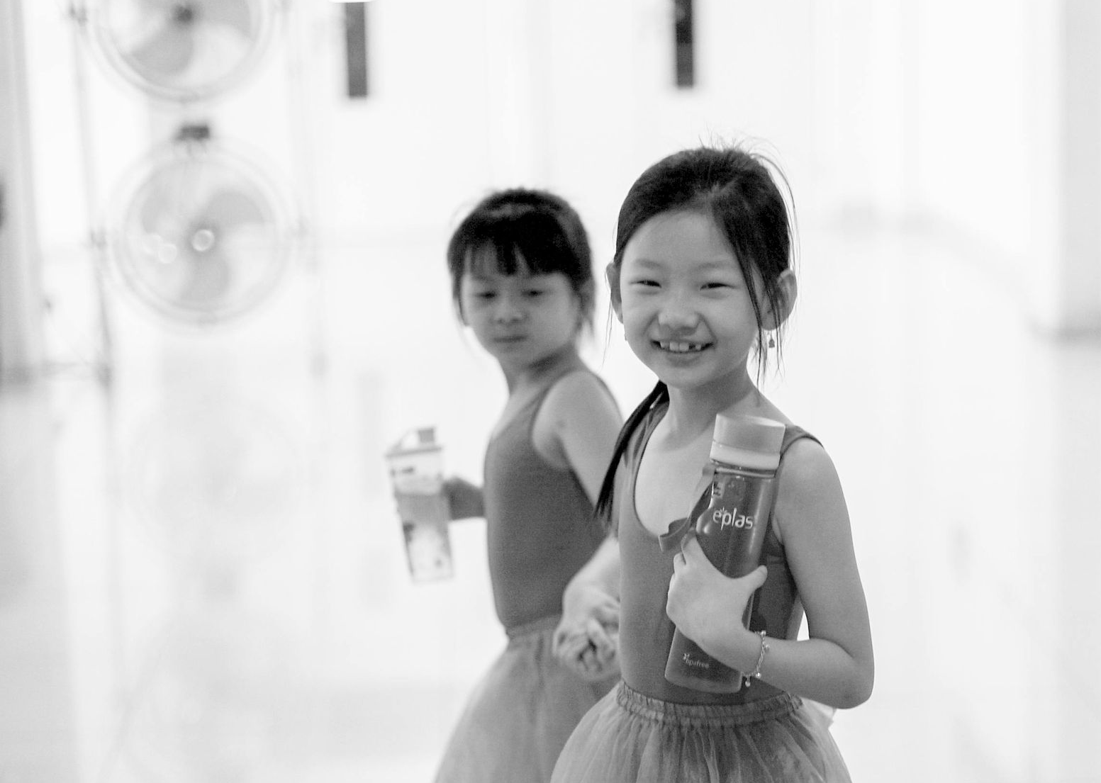 Two children in a bright hallway, one smiling and holding a bottle, the other standing behind her