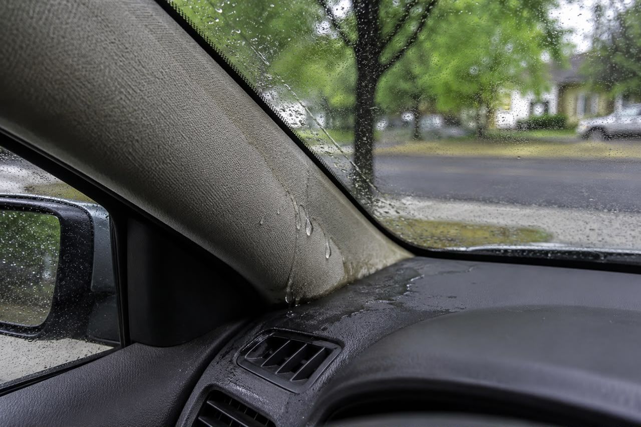 Car interior with a rain-speckled windshield and side mirror, looking out at trees and a wet street