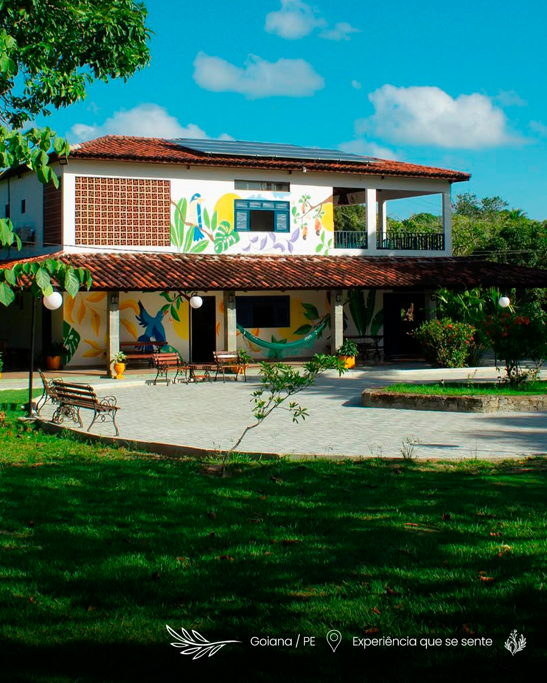 Casa de dois andares com telhado de terracota, paredes brancas e gramado verde sob um céu azul brilhante.