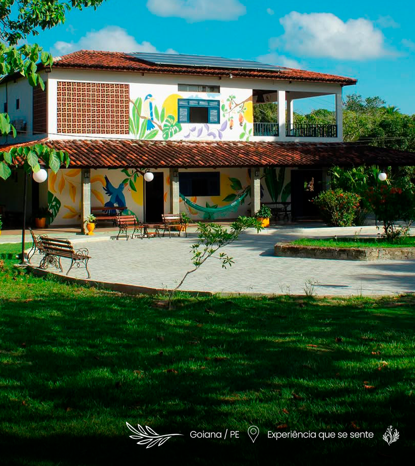 Prédio de tijolos de dois andares com mural colorido, telhado de telhas vermelhas e pátio sombreado em um jardim verdejante.
