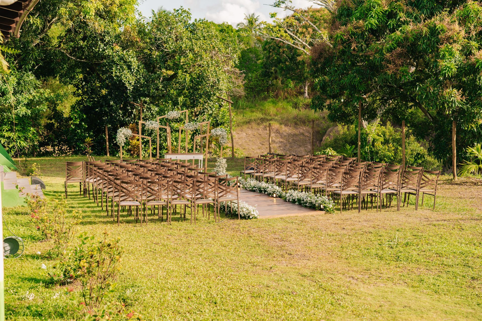 Cerimônia de casamento ao ar livre com fileiras de cadeiras na grama ao lado de um corredor branco em uma clareira na mata.