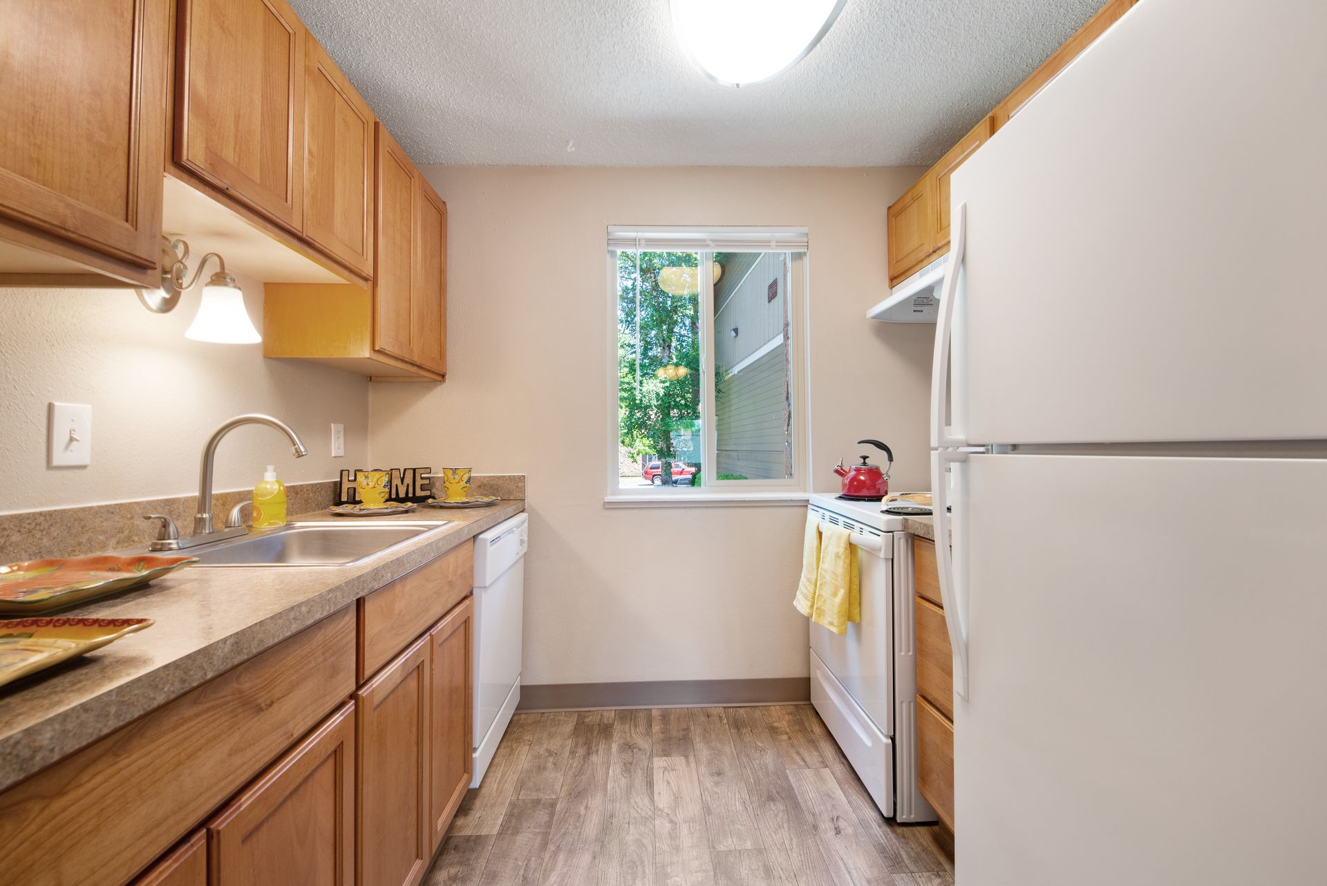 Narrow kitchen with wood cabinets, white appliances, beige counters, and a small window at the end.
