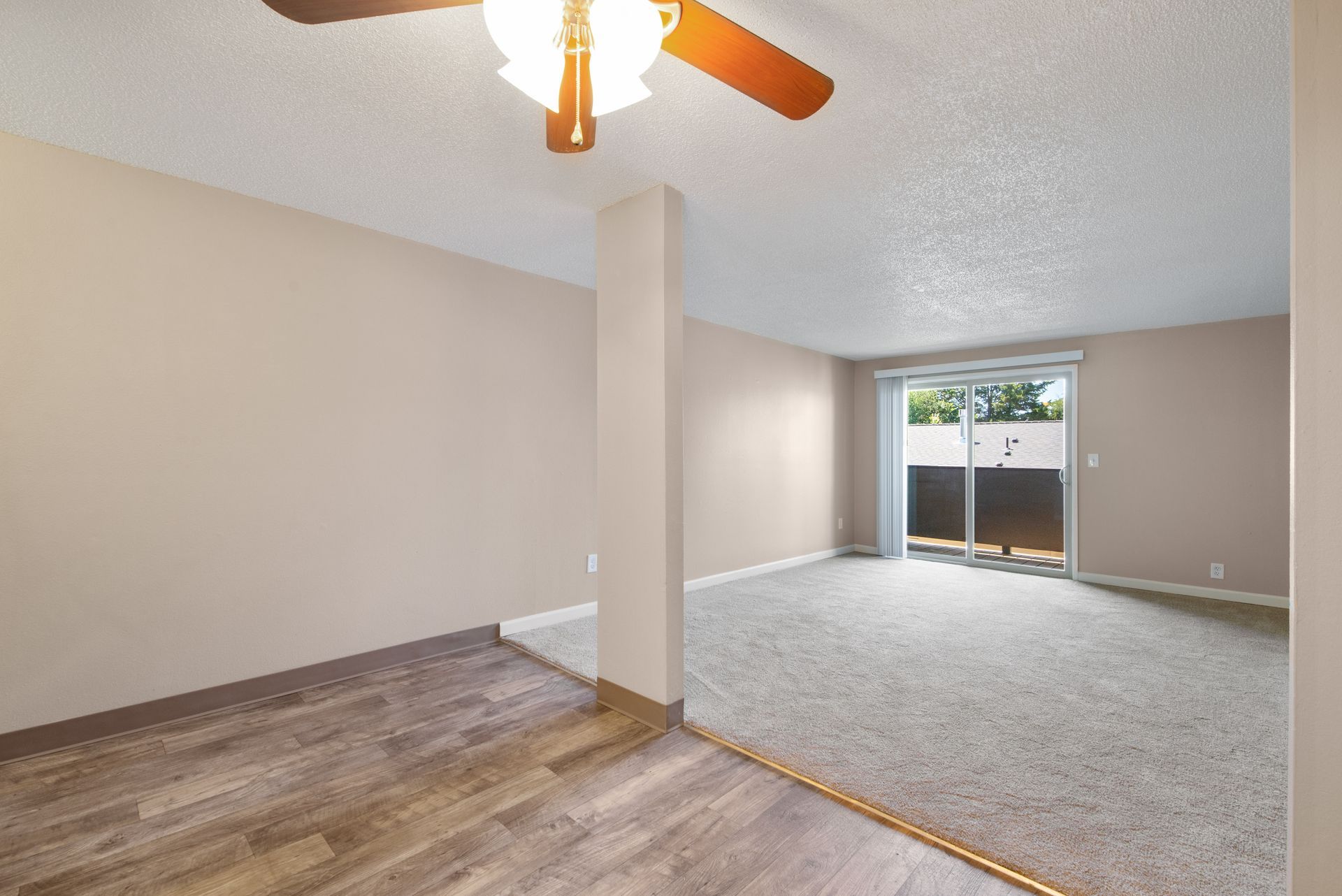 Empty living room with beige walls, wood-look flooring, carpeted area, ceiling fan, and sliding glass door.