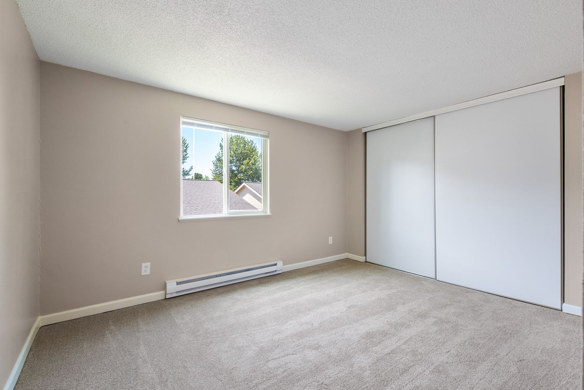 Empty beige bedroom with carpet, window, baseboard heater, and sliding closet doors