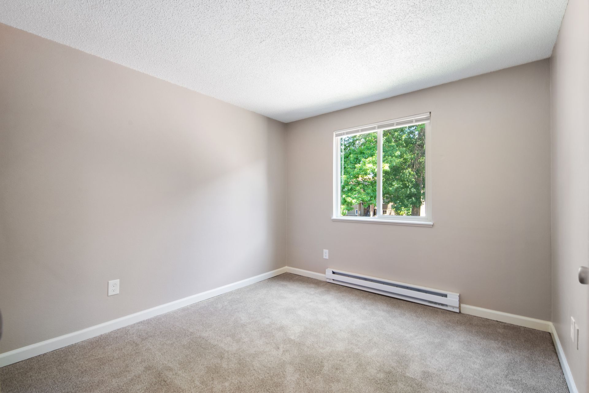 Empty beige bedroom with carpet, white trim, and a small window above a wall heater.