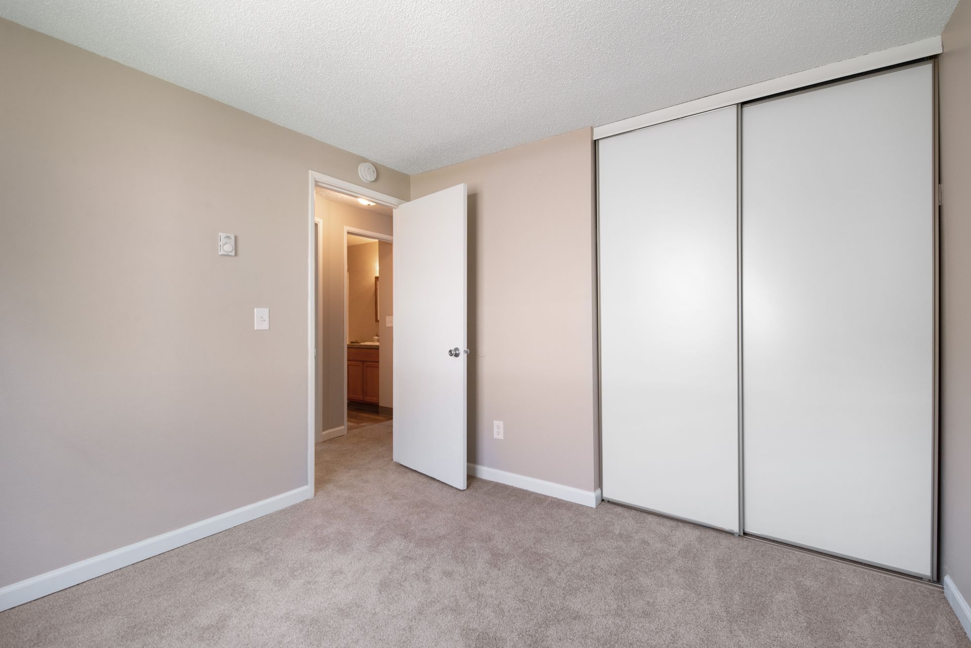 Empty beige bedroom with carpet, open door to hallway, and white sliding closet doors