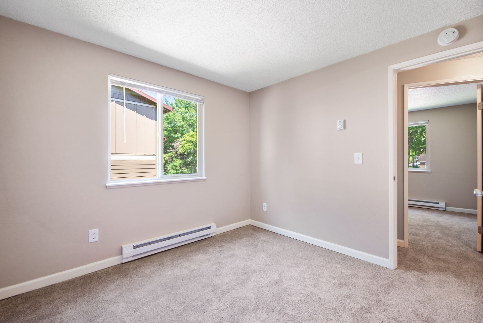 Empty beige bedroom with carpet, a window, wall heater, and doorway to a bathroom.