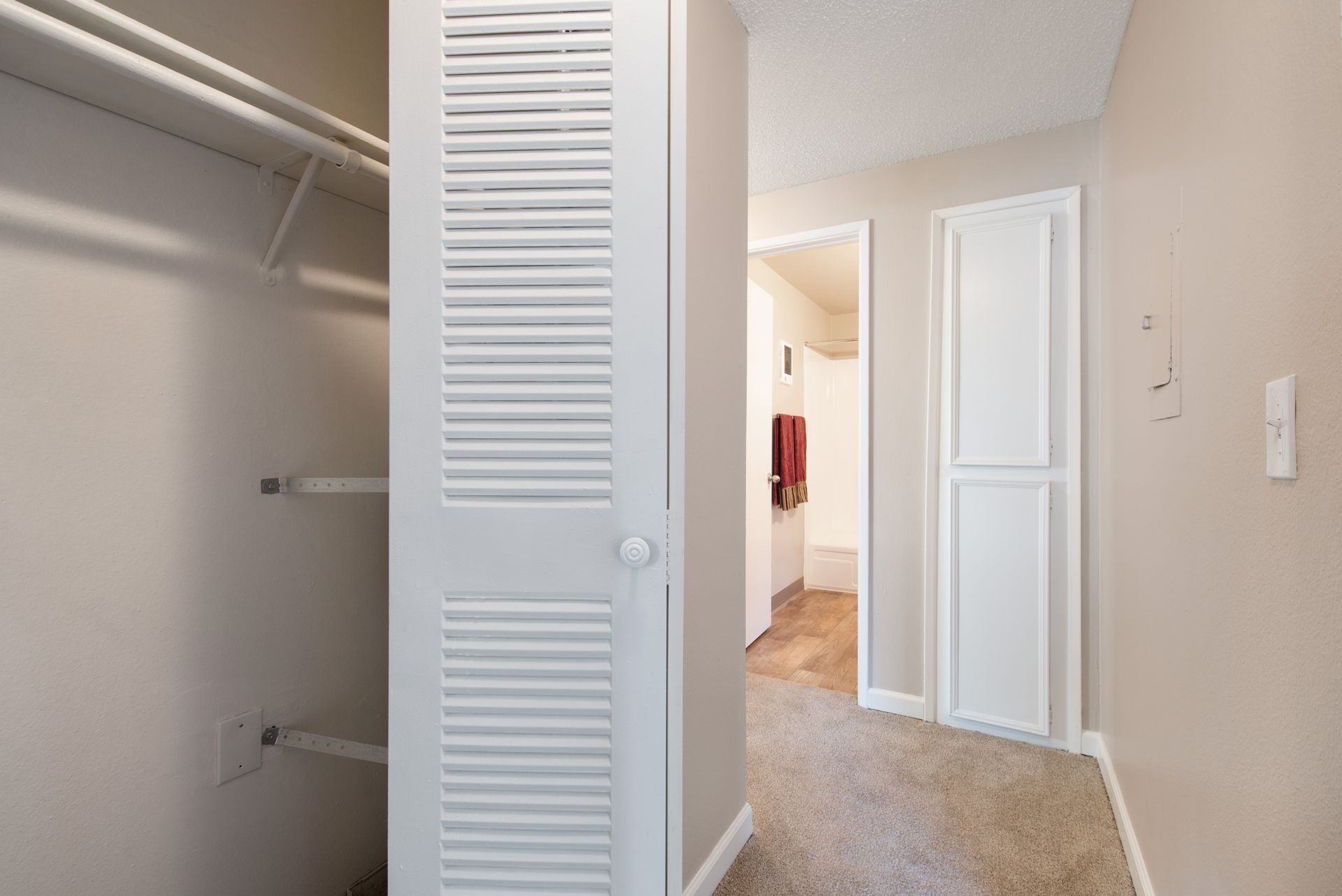 Empty closet with white louvered doors and shelves, opening to a carpeted hallway.
