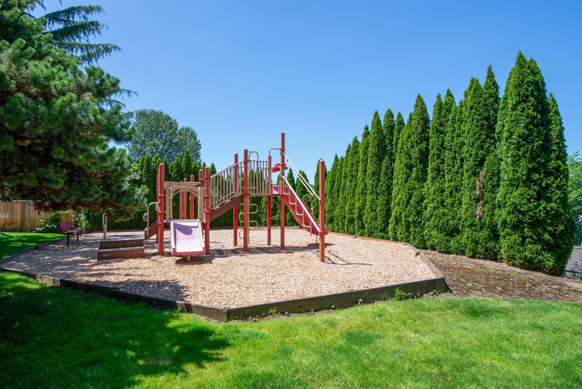 Playground with red climbing structures and slide on a woodchip area, surrounded by green grass and tall evergreens