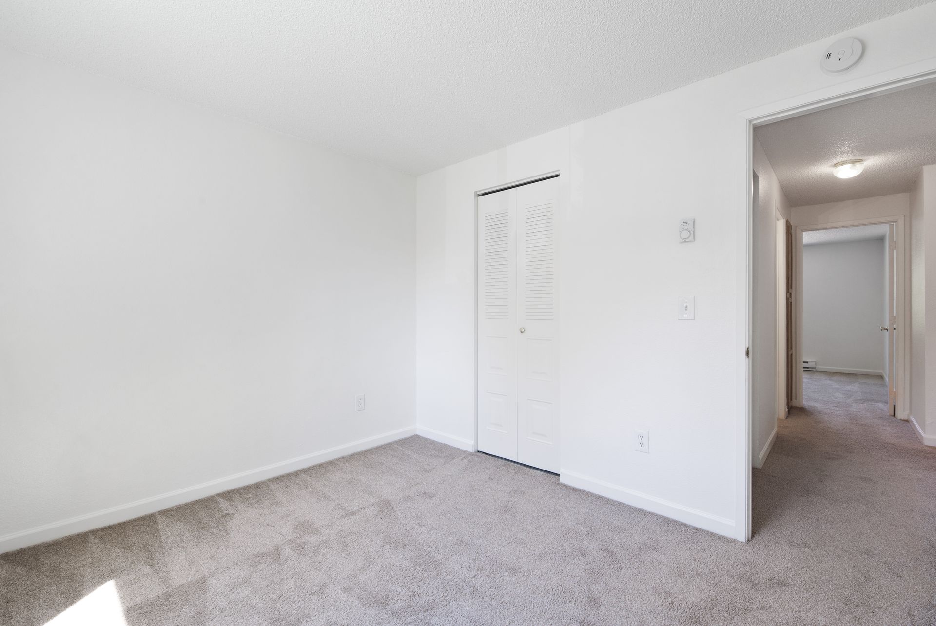 Empty white bedroom with carpeted floor and closet door, viewed from the corner near a hallway.