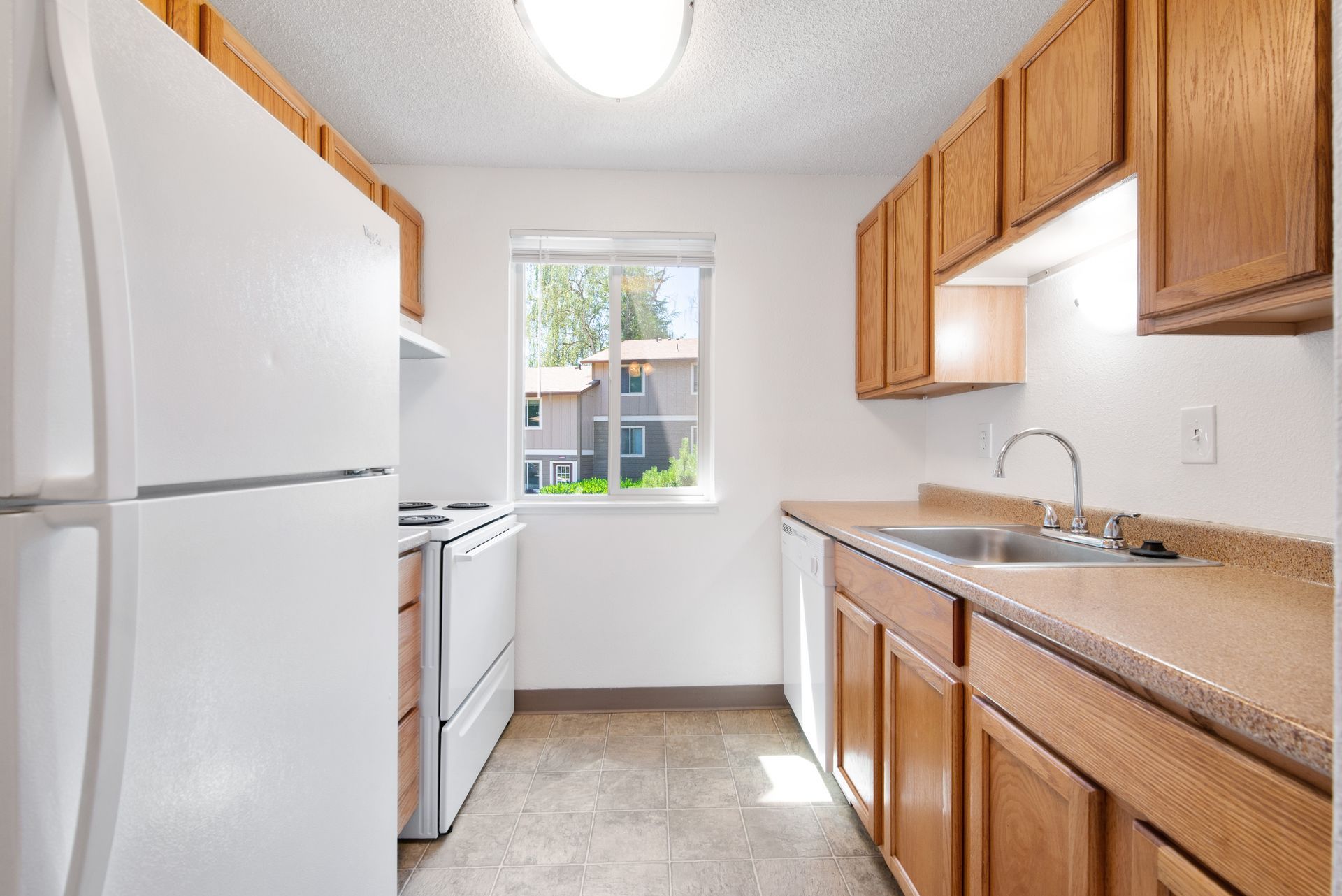 Narrow kitchen with white appliances, wooden cabinets, and a window at the end.