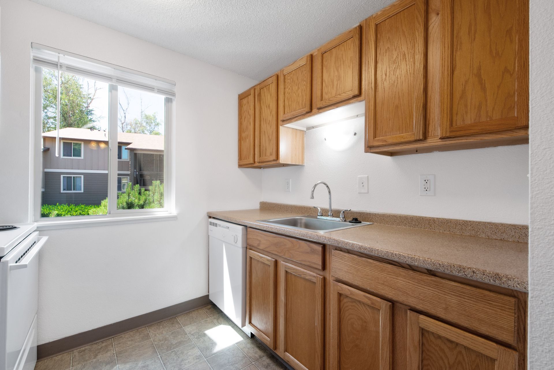 Bright empty kitchen with wooden cabinets, beige counters, sink, and a window overlooking neighboring houses