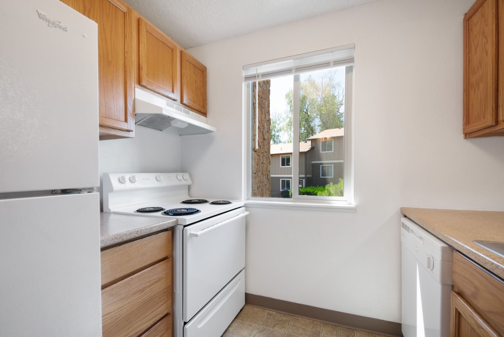 Small white kitchen with stove, refrigerator, wooden cabinets, and a window over the sink.