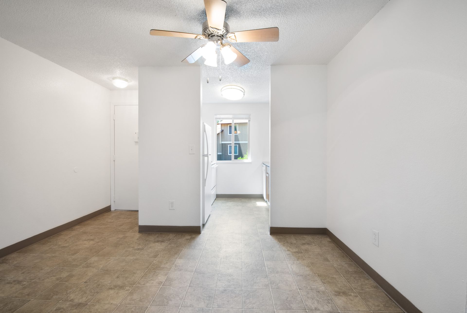 Empty white room with ceiling fan, brown flooring, and a doorway to a bright windowed area