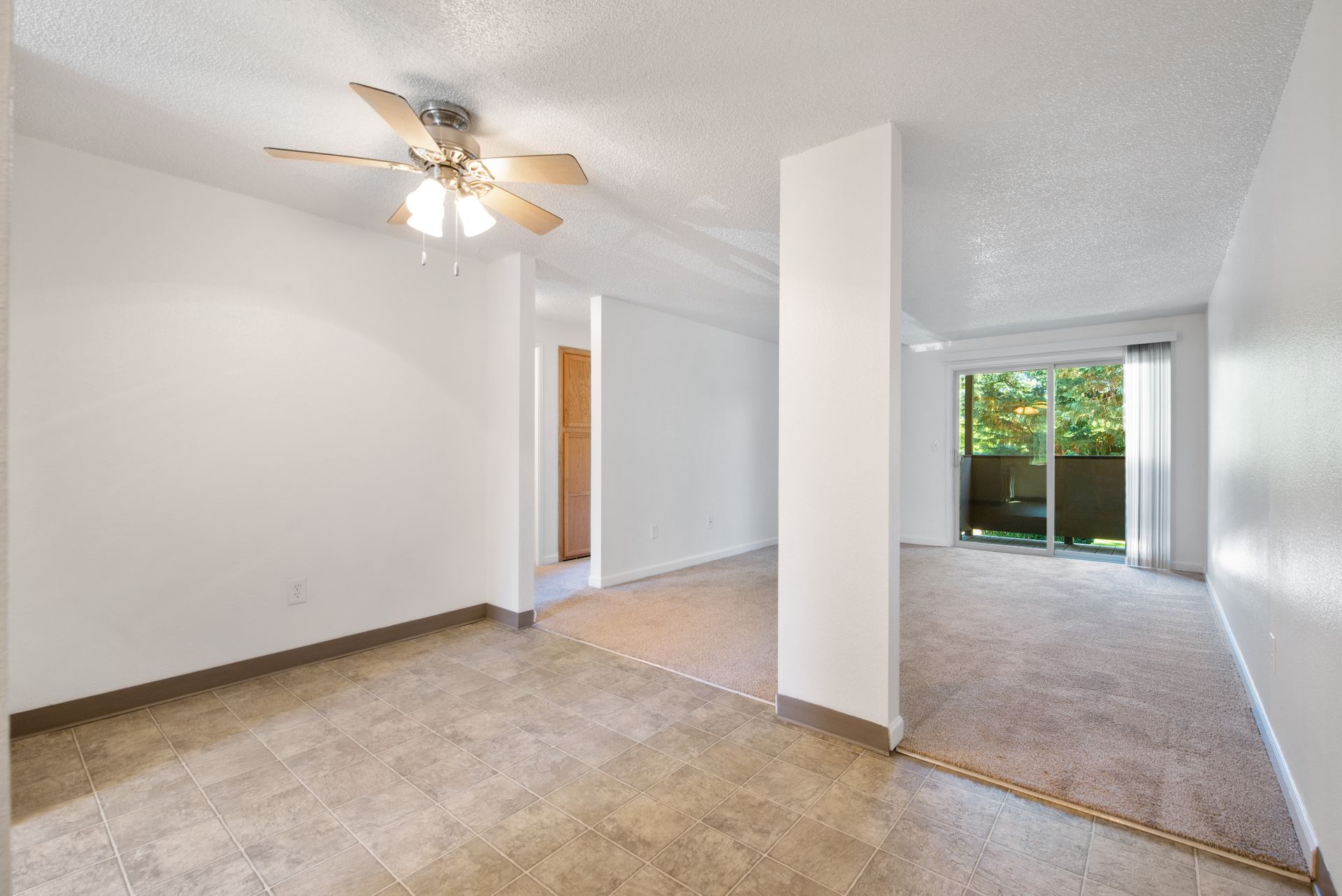Empty apartment interior with beige carpet, white walls, ceiling fan, and sliding glass door to greenery