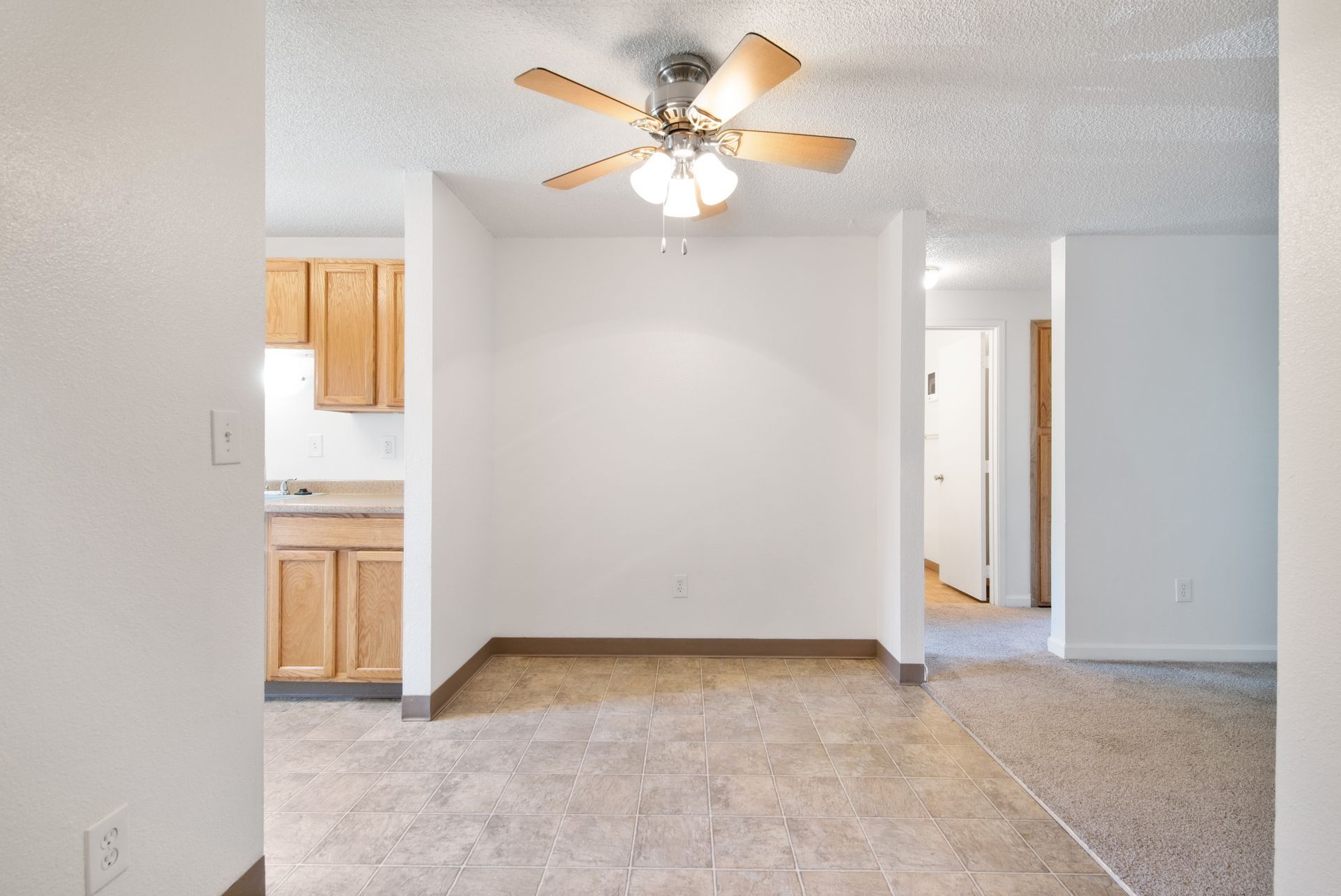Empty apartment room with tile floor, ceiling fan, and view into kitchen and hallway