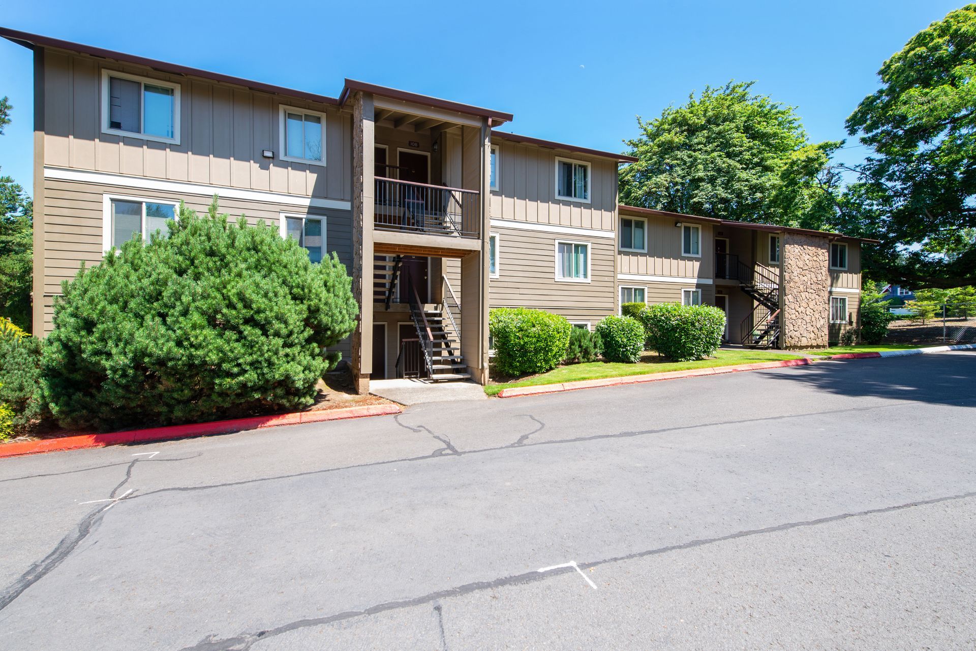 Apartment building exterior with tan siding, green bushes, and a parking lot in front