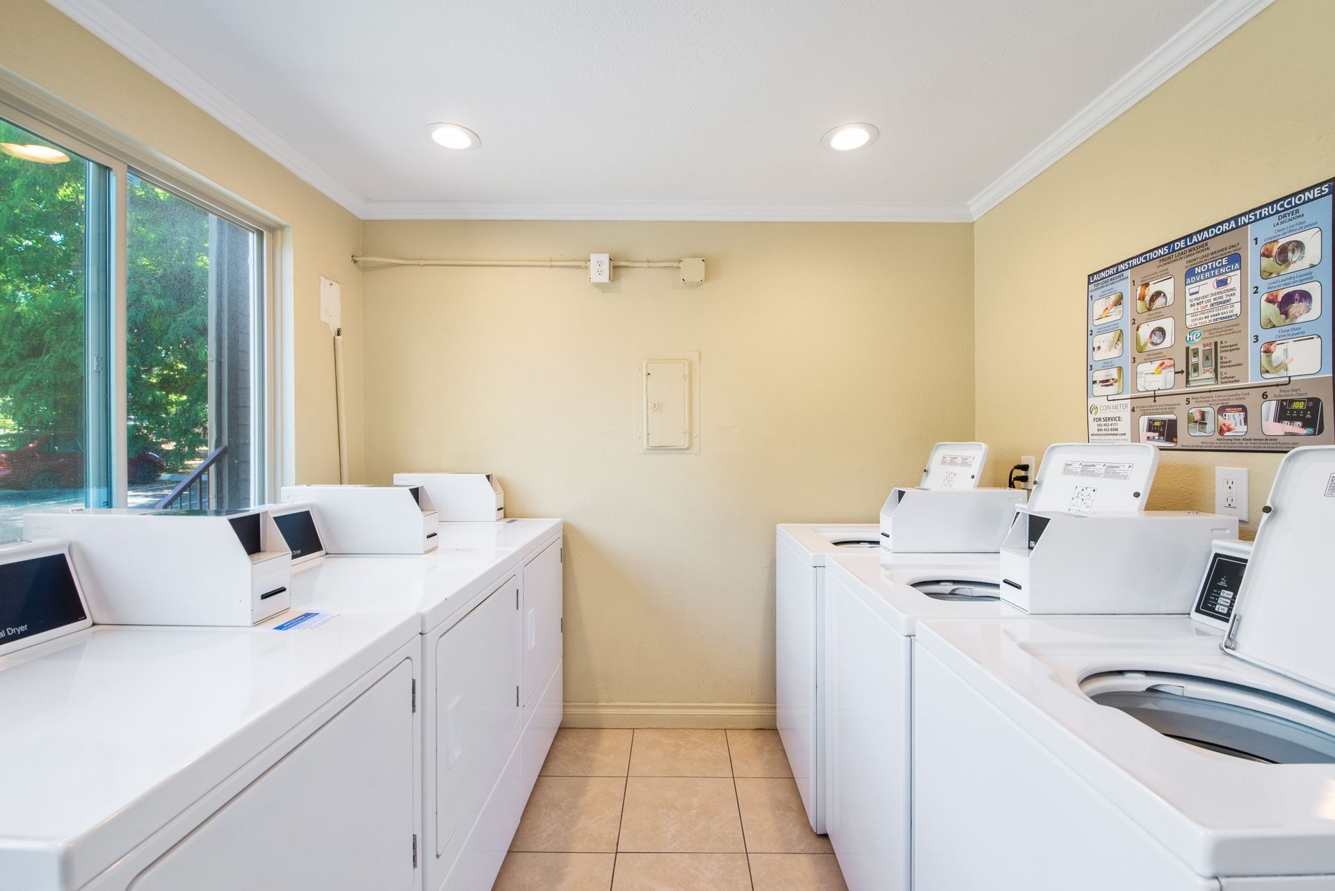 Laundry room with rows of white washers and dryers along beige walls and a window on the left