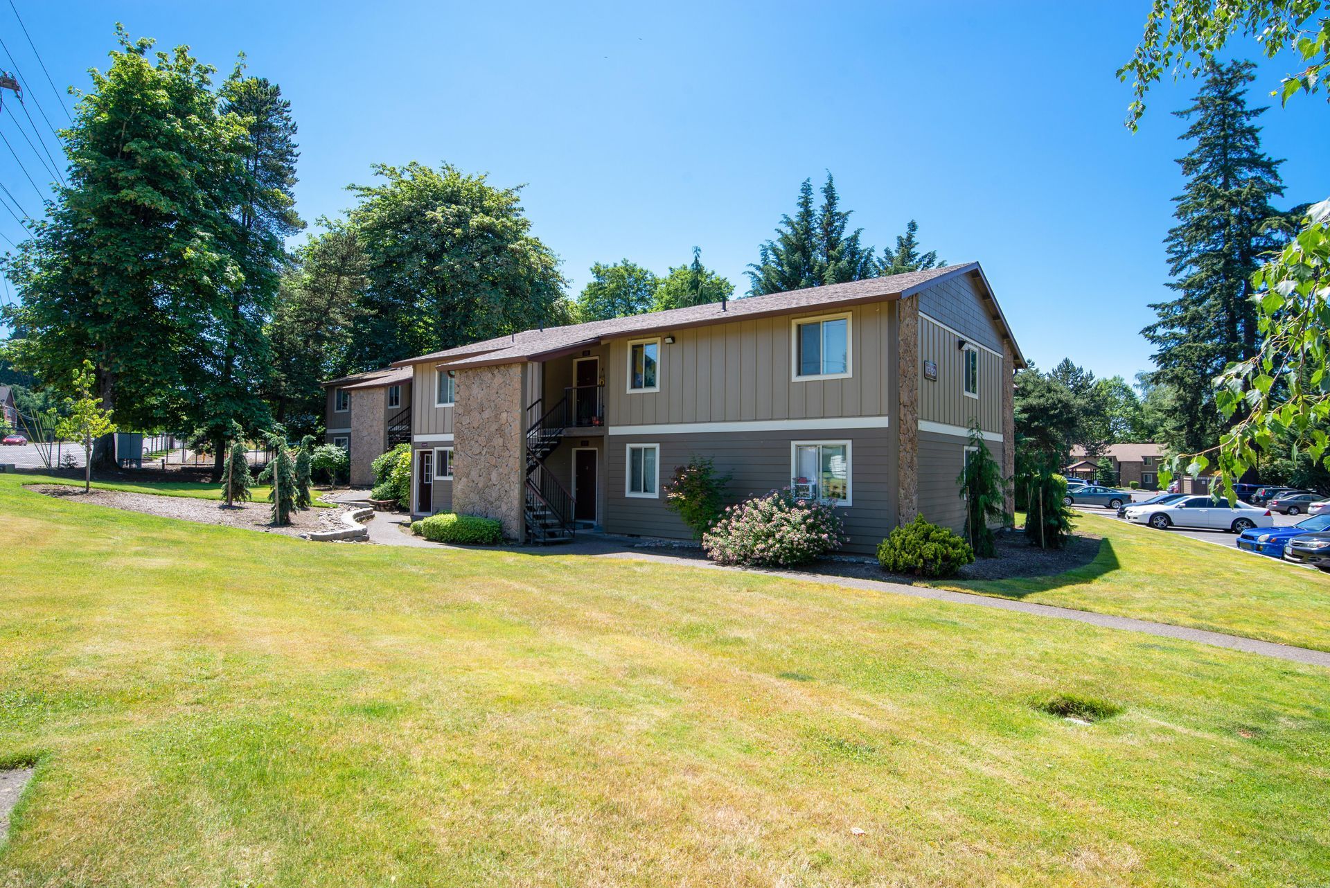 Suburban two-story apartment building with brown siding, surrounded by grass and trees on a sunny day