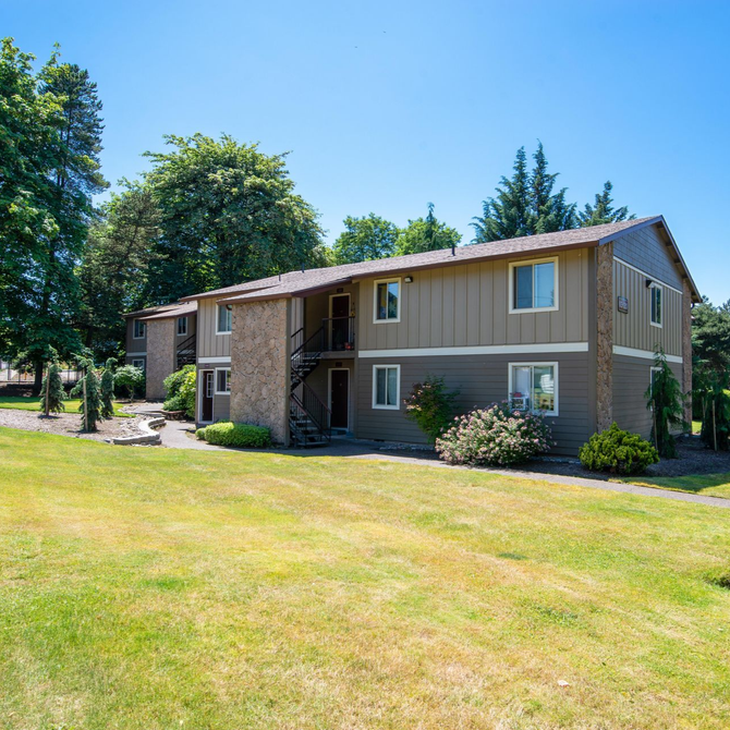 Two-story tan apartment building with stone accents, surrounded by grass and trees on a sunny day