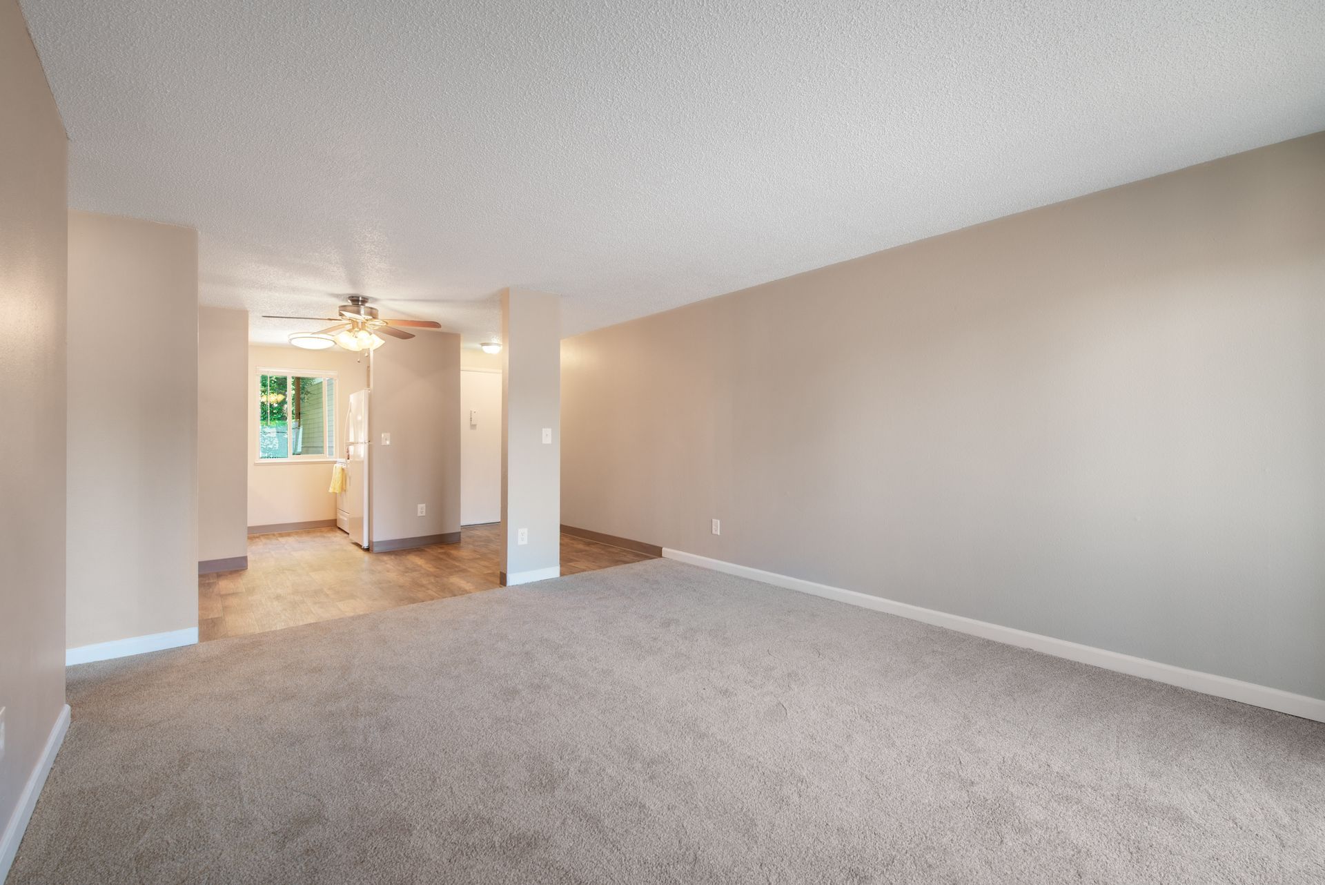 Empty beige carpeted living room with white walls and an open kitchen area