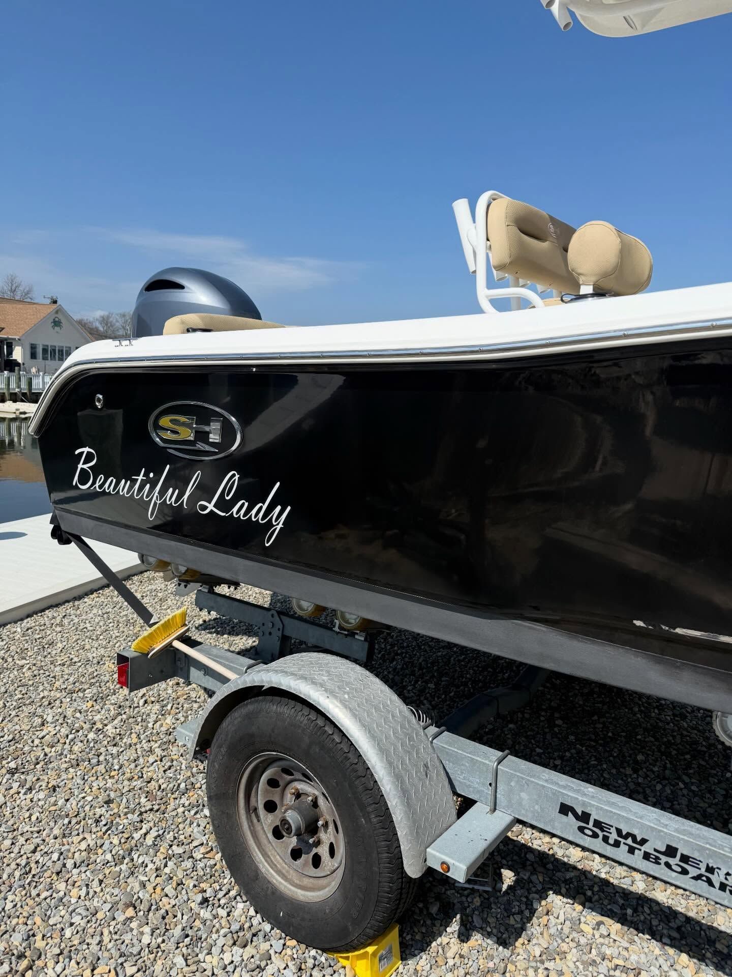 Black boat on a trailer with “Goddess of Lady” lettering, parked on gravel under a blue sky