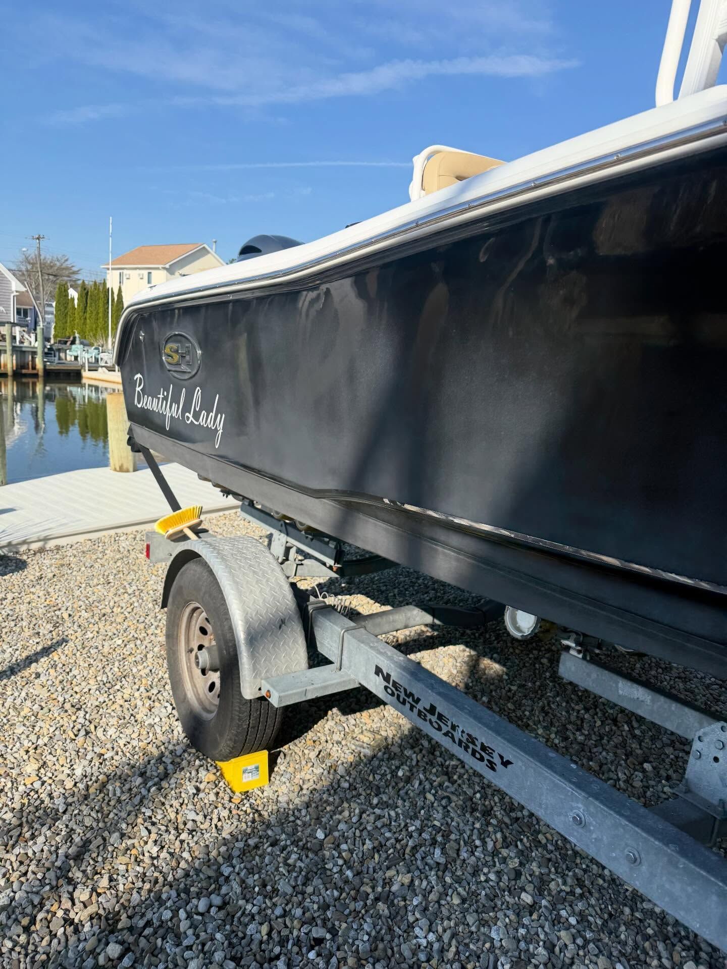 Black boat on a trailer beside a canal on a gravel lot under a blue sky