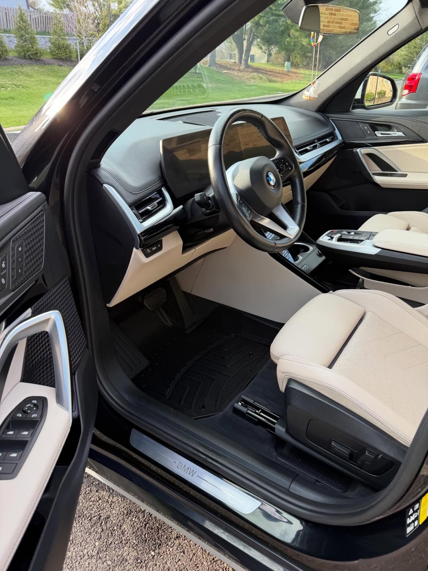 Open car door showing a beige leather interior and steering wheel, with the dashboard and driver’s seat visible.
