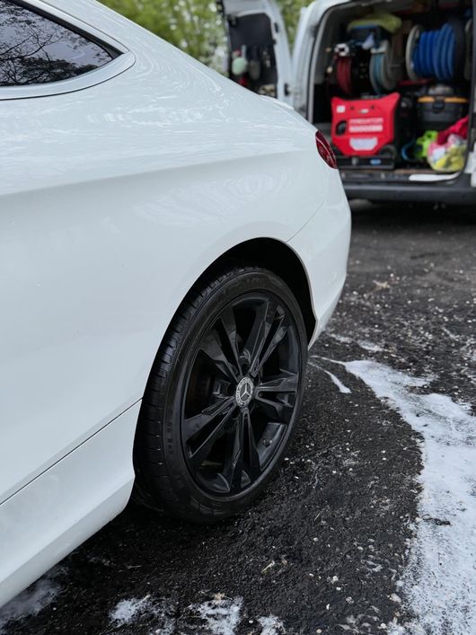 White car with black wheel parked on a wet driveway beside an open garage.