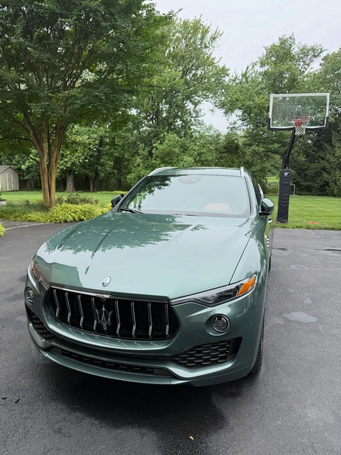 Green sedan parked on a driveway with trees and a basketball hoop in the background.