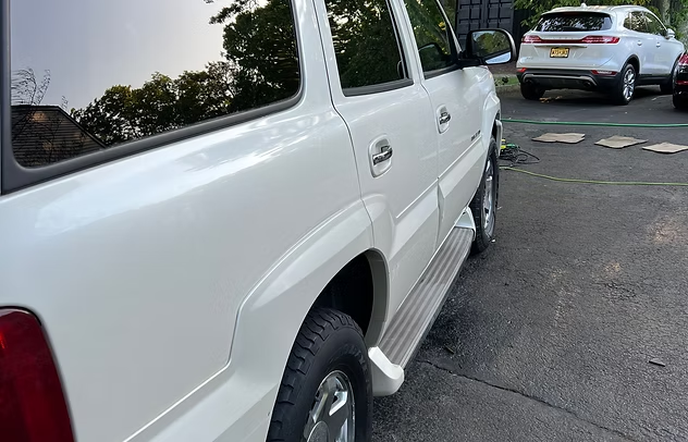 White SUV parked on a paved street beside a low wall and trees, with another car ahead.